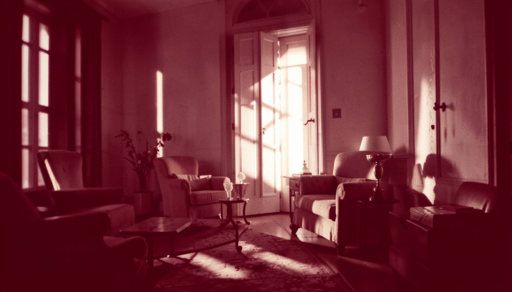 A warm duotone photograph in deep maroon and cream tones, showing an atmospheric interior with soft natural light and elegant shadows.